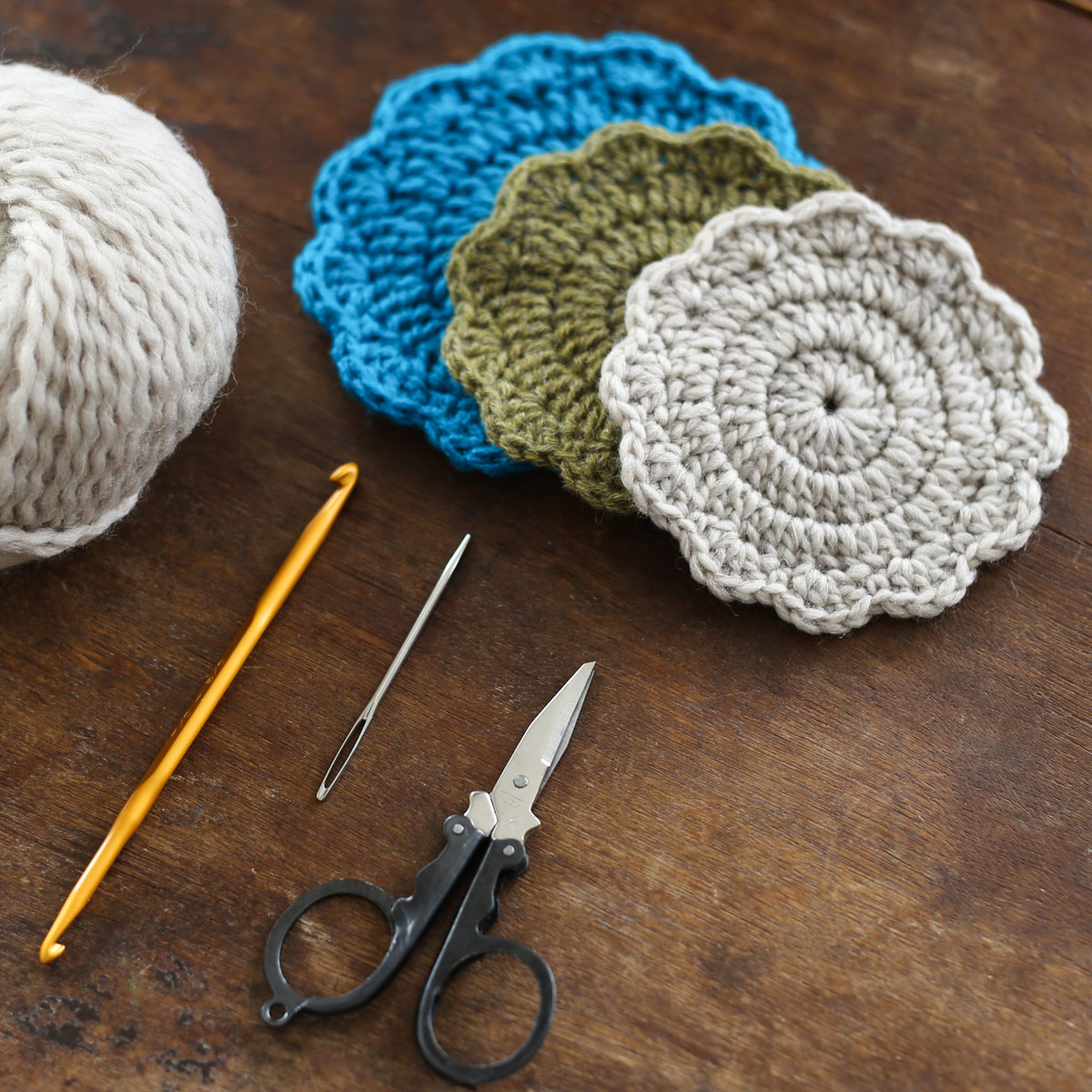 定番小物からはじめる かぎ針編み初心者コース［第1回］。長編みで編む輪編みコースター。完成作品3色と、必要な材料（毛糸玉、かぎ針、とじ針、ハサミ）が木製のテーブルに並んでいる様子。