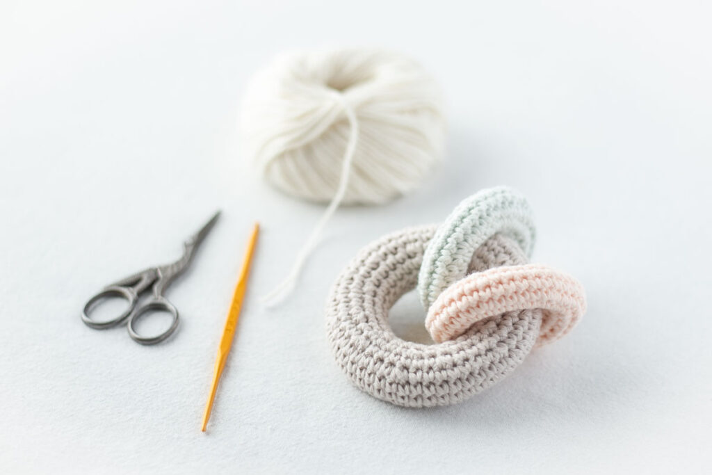 パステルカラーのコットン糸で編んでいるかぎ針編みのラトル（赤ちゃんのガラガラ）。白い毛糸とかぎ針、ハサミも並んだ製作中の様子。