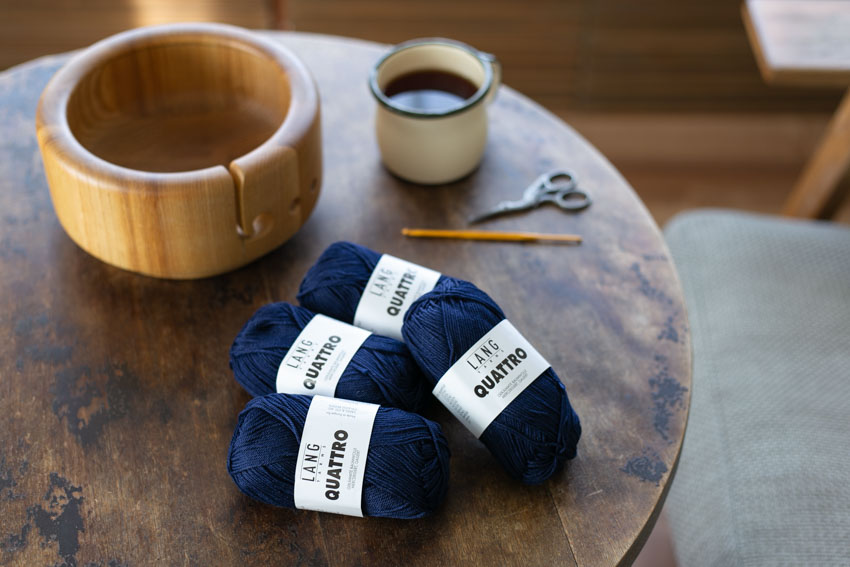 かぎ針編みのある風景。テーブルにはかぎ針とハサミ、ヤーンボウル。編み物の原動力になるエレガントなネイビーのコットン糸。